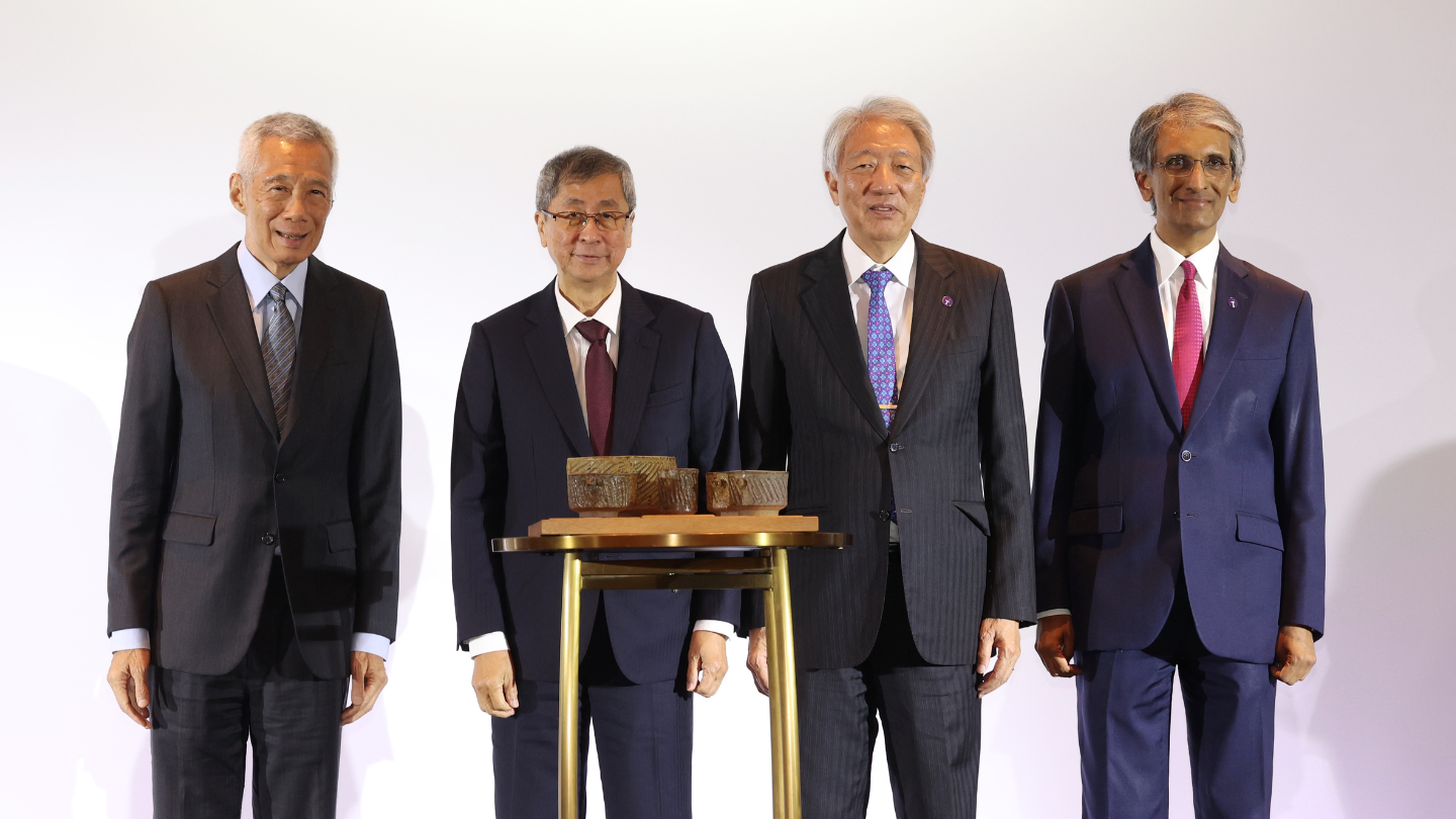 Four men in suits pose beside a small artifact display on a pedestal, white backdrop.